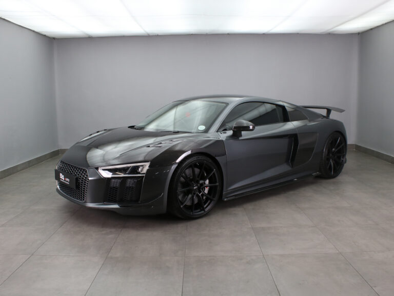 Dark gray Audi R8 sports car in a bright showroom, shown in a front three-quarter view with black wheels and rear spoiler.