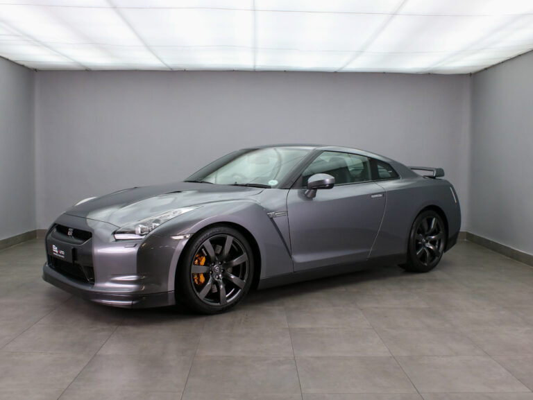 Front three-quarter view of a metallic gray Nissan GT-R sports car in a clean showroom, showing gold calipers and dark alloy wheels in detail.