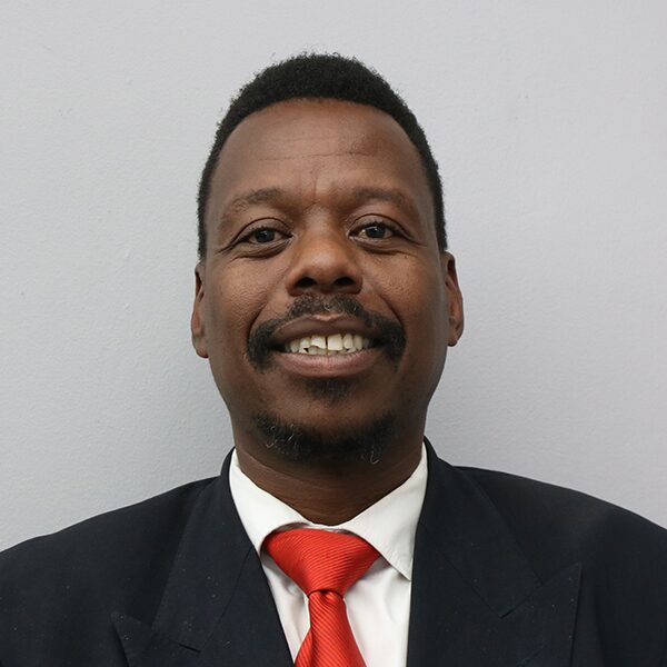 Portrait of a man in a dark suit and red tie, smiling at the camera against a light gray background.