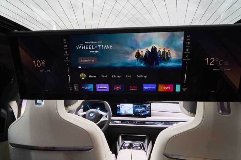Inside a luxury car cabin with a wide horizontal infotainment screen displaying a 'The Wheel of Time' banner and app icons. Beige seats surround a dark dashboard.